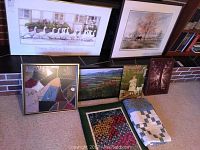 Seven framed art pieces including watercolor print signed by Dawna Barton, and two folded quilts placed on floor against wall.