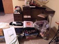 Corner sewing table with cushioned stool and various sewing supplies including fabrics, patterns, wool yarns in plastic bags, and sewing threads; wooden storage drawers and shelves containing additional items.