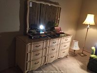 Front view of off-white dresser with nine drawers and matching mirror on top, surrounded by a floor lamp and green chair in the room.