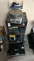 Full view of stereo stand holding stacked vintage tape reel cans, with numerous electronic devices arranged on its shelves, some with visible branding including 5 CM on the reel cans