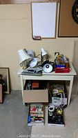 Wide shot of small desk with assorted office supplies and lamps on top plus boxes of stationery underneath, white dry erase board on wall
