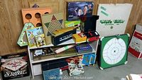 Assortment of board and card games in original boxes arranged on and around wooden shelving. Includes Scrabble, Pictionary, other card and board games, and a dry erase game board.