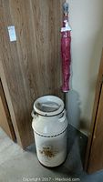 Hand painted steel milk jug with floral designs, placed beside a wrapped Canadian flag on pole against a wall.