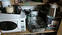 Wide view of kitchen shelf holding white Sunbeam microwave oven, multiple roasting pans with removable wire rack, and boxed Presto coffee maker.