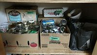 Boxes containing assorted glass canning jars, metal lids, jars with screw bands, and some packaged lids. Also visible is a metal pressure cooker.