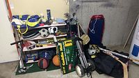 Wide shot showing various sports gear including golf clubs, basketballs, tennis rackets, hockey sticks, helmet, ski goggles, and roller skates arranged on shelves and floor.