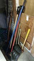 Photo showing multiple garden tools leaning against a wall including shovels, a blue rake, a black garden fork, and an axe with yellow handle.