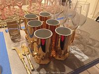 Photo showing eight shiny gold colored beer stein mugs with large handles and pedestal bases; two gold serving knives with bamboo style handles; several rose stem colored beer glasses with slender tall bowls and clear stems; small square vase.