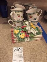 12 stacked Villeroy & Boch mugs with fruit and leaf design, beside ceramic asparagus serving bowl with decorative ribbons