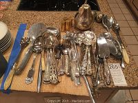 Image showing a variety of stainless steel and silver-plated serving utensils laid out on a counter including spoons, forks, pie servers, and knives with decorative handles.