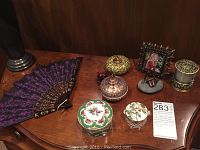 Tabletop displaying a purple and black decorative fabric fan, three ornate porcelain lidded boxes with floral designs, a figurine, picture frame, and candle holder.