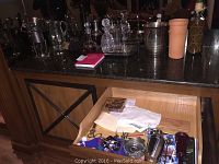 Wide view of bar countertop with multiple crystal decanters, clay wine cooler, metal ice buckets, and various bar items, plus a drawer with smaller bar accessories open beneath.