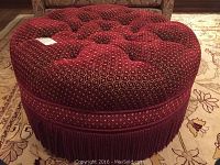 Front angled view of the round red ottoman showing the tufted top and brocade fringe trim around the bottom edge.