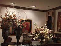 View of large decorative vases with faux plants, floral ceramic vase on gold-tone stand, and smaller floral arrangement.