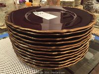 Stack of 12 maroon oversized plates with gold scalloped edges shown stacked on a table.