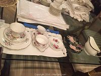 Overview of the china set on glass table with embroidered white cloth beneath, showing cups, plates, cream pitcher, and gravy bowl.