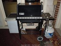 Full view of the Weber propane grill showing main cooking chamber, side burner control panel, side table, propane tank, and wheels.