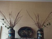 Two large black Asian themed ceramic vases filled with faux dried floral arrangements, displayed with an oval metal tray painted with birds and fruit, all on top of a table runner.