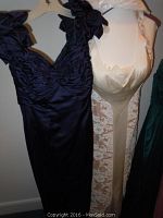 Two evening gowns hanging side by side, navy Vicky Tiel gown with floral shoulder decorations and white satin brocade gown with lace details