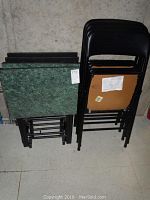 Stack of four TV trays with green faux marble tops and folding black metal legs beside a stack of four black folding chairs with padded seats and backs.