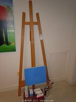 Tall natural wood easel with blue canvas board and several painting supplies including bottles, brushes in container, and palette paper on floor