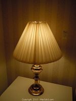 Full view of one brass table lamp with pleated cream-colored fabric shade illuminated.