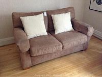Front view of brown suede-like fabric love seat with two light-colored throw pillows, placed against wall on wooden floor.