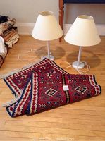 Pair of white wood table lamps with white fabric shades positioned behind a folded wool rug with red and blue geometric pattern on wooden floor.
