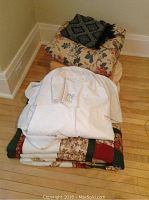 View of linen lot stacked on floor showing bed skirt, black patterned throw on top, floral quilt, wool blankets, and patchwork quilt underneath.