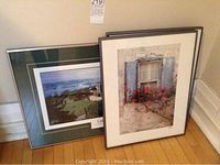 Two framed photographic prints leaning against a wall. One print is a panoramic view of a castle and landscape, the other is an image of a window with flowers.