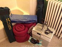 Items including two rolled red sleeping bags, two Wild Country camping chairs in carrying bags, a blue tarp, an electric cooler, a headlamp, a flexible mini tripod, binoculars, and a small folding chair with striped fabric.