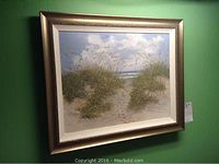 Framed painting hanging on wall showing full composition of dunes, grasses, ocean and sky.