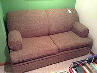 Brown loveseat fold out bed situated in a corner with a green and beige wall. The loveseat has textured brown fabric upholstery and matching cylindrical arm cushions.