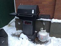 Black Vermont propane BBQ grill with cover, propane tank, and two side shelves, set outside in snow.
