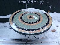 Top view of the round outdoor table showing the mosaic tile design with snow on top.