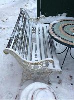 Side view of bench showing cast iron frame with scroll and floral motifs, wood slat seat and backrest covered with light snow.