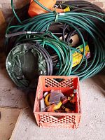 Multiple coiled green garden hoses with some black hoses and a collapsible green hose lying on the ground beside an orange plastic crate filled with hose attachments.