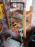 Manual green push reel lawn mower standing in front of a wooden shelf with various garden items and equipment nearby.