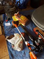 Full lot overview showing shop vac, bags, orange weed wacker, and black leaf blower on blue tarp.