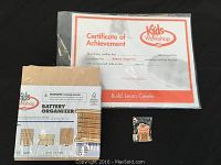 Photo showing the battery organizer craft kit wrapped in plastic, the achievement certificate, and a small metal completion pin.