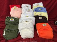 Photo shows 17 cloth diapers in multiple colors arranged on red fabric backdrop. Diapers have labels indicating brand Anne Marie Padorie, various colors including green, orange, red, pink, white, brown, blue, yellow. Velcro closures visible on some.