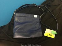 Photo shows a blue leather crossover purse lying flat on a black cloth or fabric background, with an adjustable strap and a small tag attached.