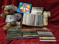 Photo showing the entire lot arranged on red fabric background. Includes stacked CDs, plush toy dressed in religious armor, and stainless steel water bottle with printed design.