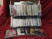 Two stacks of approximately 80 assorted new CDs in original packaging shown on red cloth background. CDs are in jewel cases with cover art mostly visible.