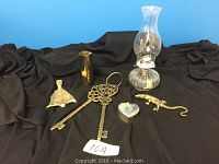 Photo showing a group of five items: brass vase, brass salamander figurine, brass fish dish, glass oil lamp with chimney and wick, and a small silver plated heart-shaped box with red velvet interior on a black fabric background.