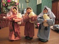 Three Byers Choice caroler figurines on a wooden table in a traditional-style room. Each holds a white lamb and wears fabric robes and headscarves.