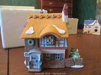 Front and left side view showing yellow thatched roof with snow patches, small blue door with sign, wreath decoration, chimney and snowy base with small trees.