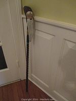 Full view of wooden walking stick leaning against white wall and door showing carved and painted Santa Claus head handle and textured shaft.