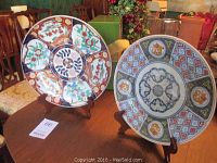Both charger plates displayed upright on wooden stands on a wooden table with surrounding furniture and decor visible in the background.