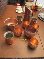Full set of assorted copper vessels arranged on a wooden table including bowls, mugs, cups, a pitcher, and a small handled pot.